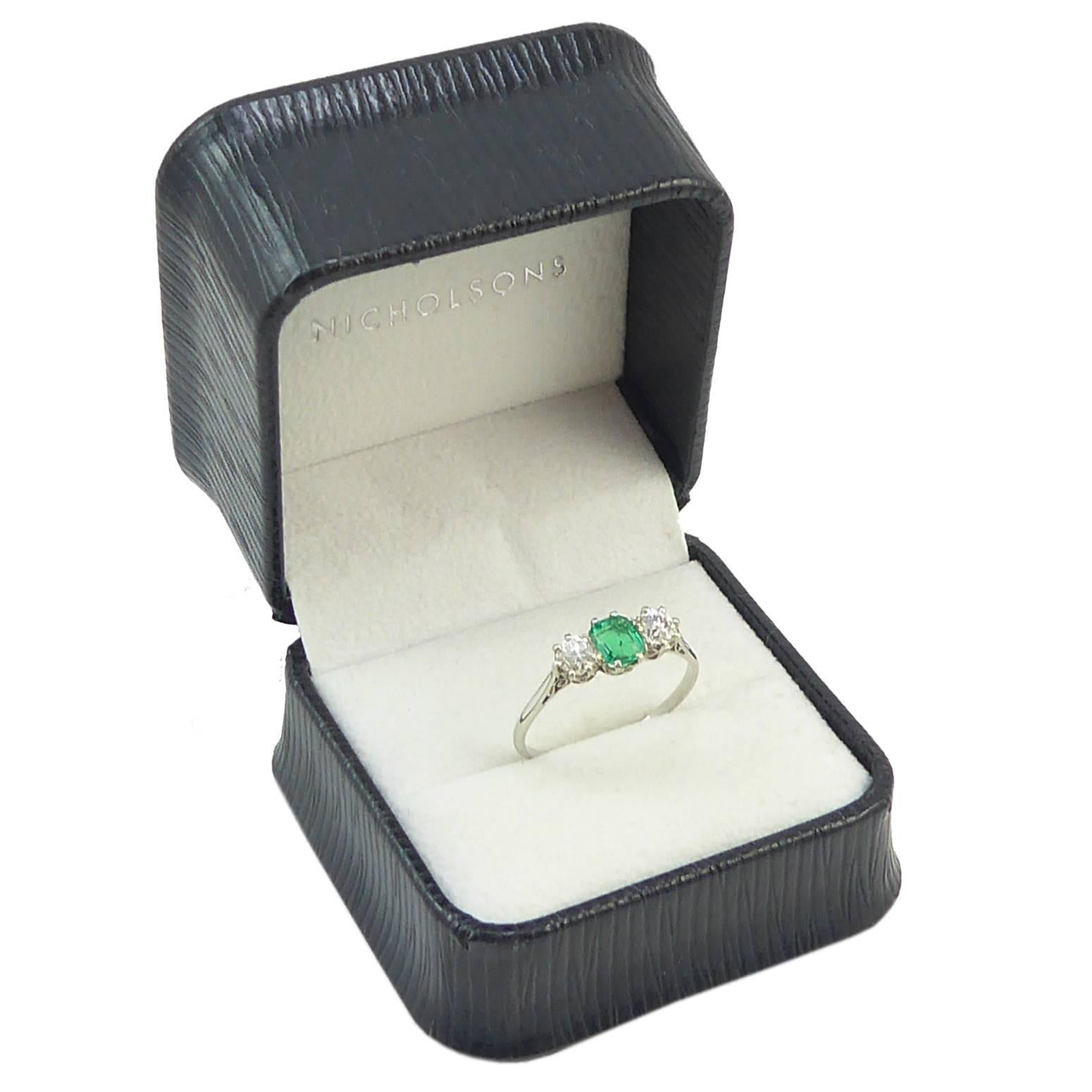 A classic three stone emerald and diamond engagement ring set to the centre with a lovely bright green coloured emerald, octagonal in shape, flanked to either side with an old European cut diamond.

All the gemstones are claw set in white metal to a