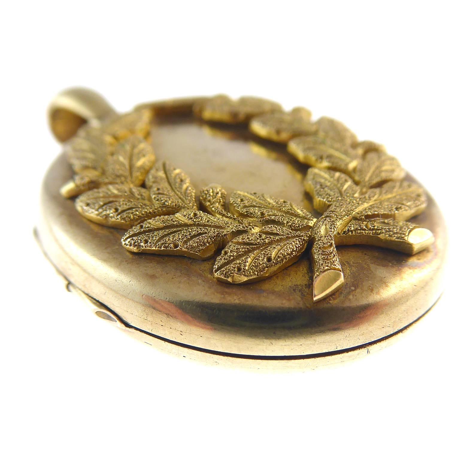 An oval hinged locket measuring approx. 28mm x 35mm with applied engraved wreath shaped panel and a plain polished back.  The front depicts a wreath of acorns and oak leaves with a beautifully burnished gold colour.  The locket display and overall