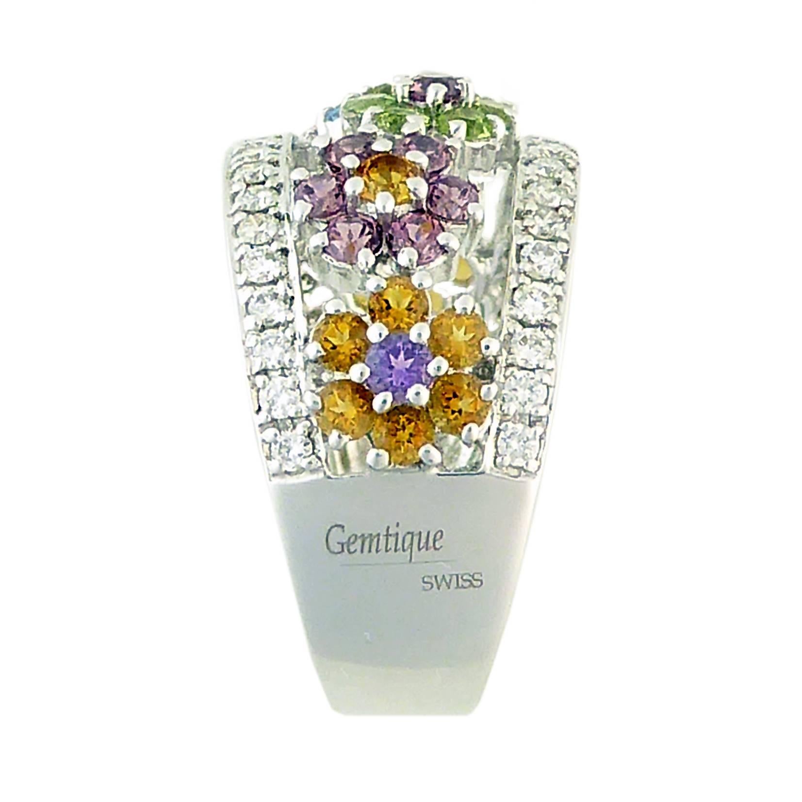 An extraordinarily delightful ring featuring a rainbow of colourful flowers set with an assortment of gemstones in a row across the finger.  To each side of the floral row is a line of channel and bead set sparkling brilliant cut diamonds, approx.