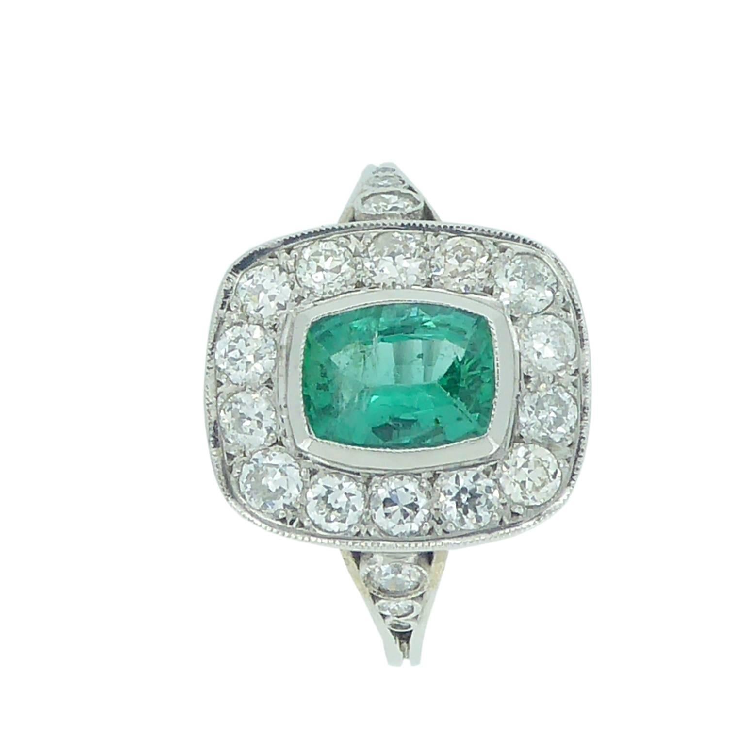 A lovely cushion shaped emerald of good green colour sits in a halo of brilliant cut diamonds, the whole arrangement reminiscent of the Art Deco era.  
This pre-owned cluster ring is perfect as a dress or engagement ring and the diamond decorated