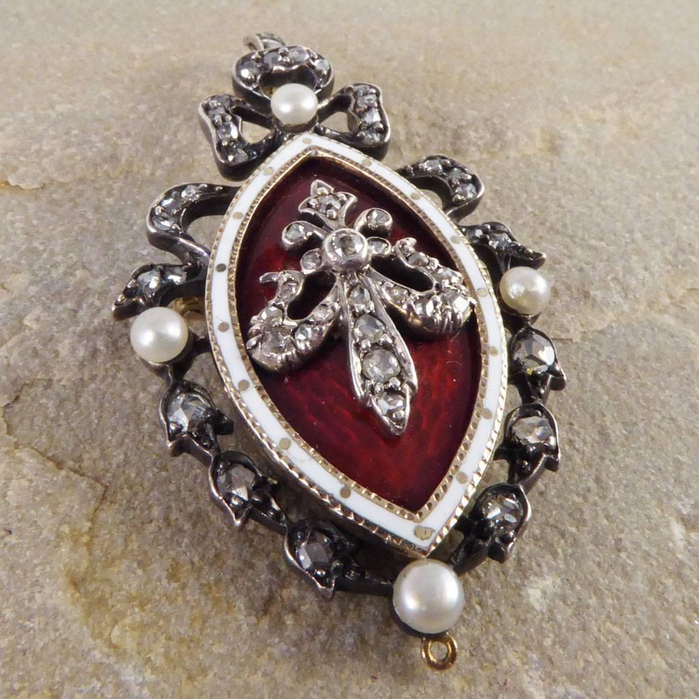 This magnificent antique brooch pendant was crafted in the Victorian era. With blood red and white enamel decoration set in gold silver, this piece features a wonderful diamond and pearl set ribbon surround. 

Wear as a brooch or pendant, this