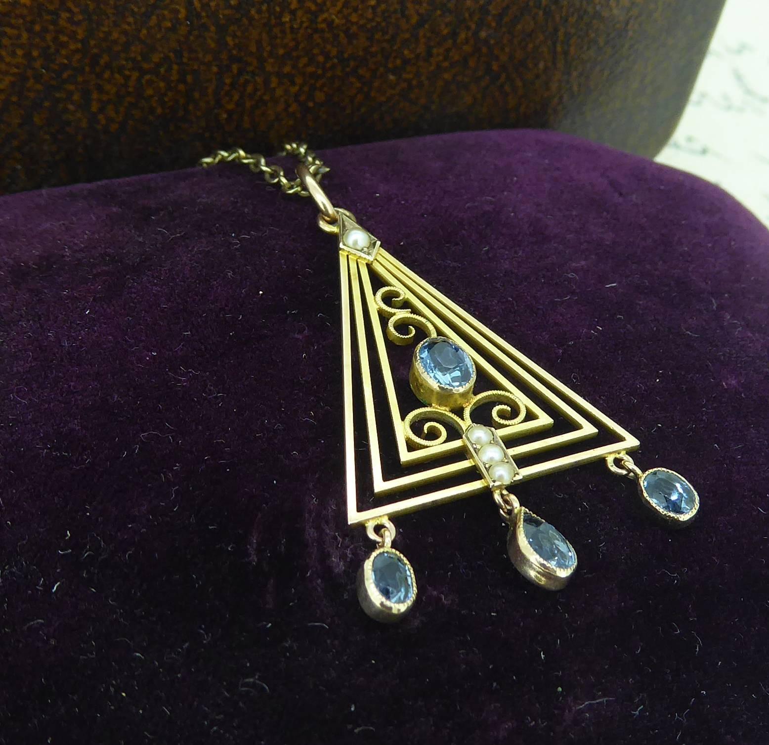 A delicious Art Deco pendant in a typical geometric/pyramid shape reminiscent of the Egyptian influence of the era.  Centrally set with an oval mixed cut aquamarine in a rub over setting with millegrain edged decoration and with gold wire curclicue