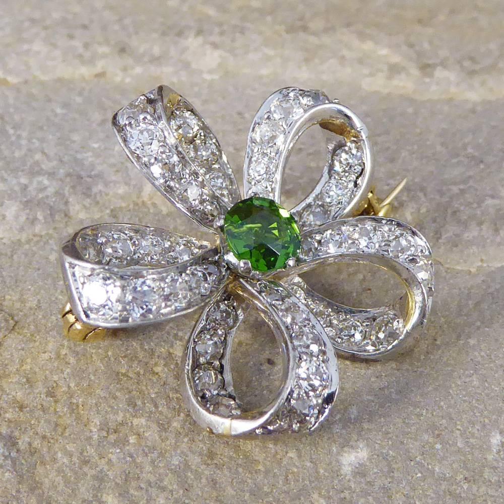 This pretty Late Victorian brooch can also be used as a pendant with a hinged loop on the back. Showcasing one Green Garnet stone weighing 0.44ct supported by five Diamond set ribbons creating a flower aesthetic. With 45 sparkling Diamonds adorning
