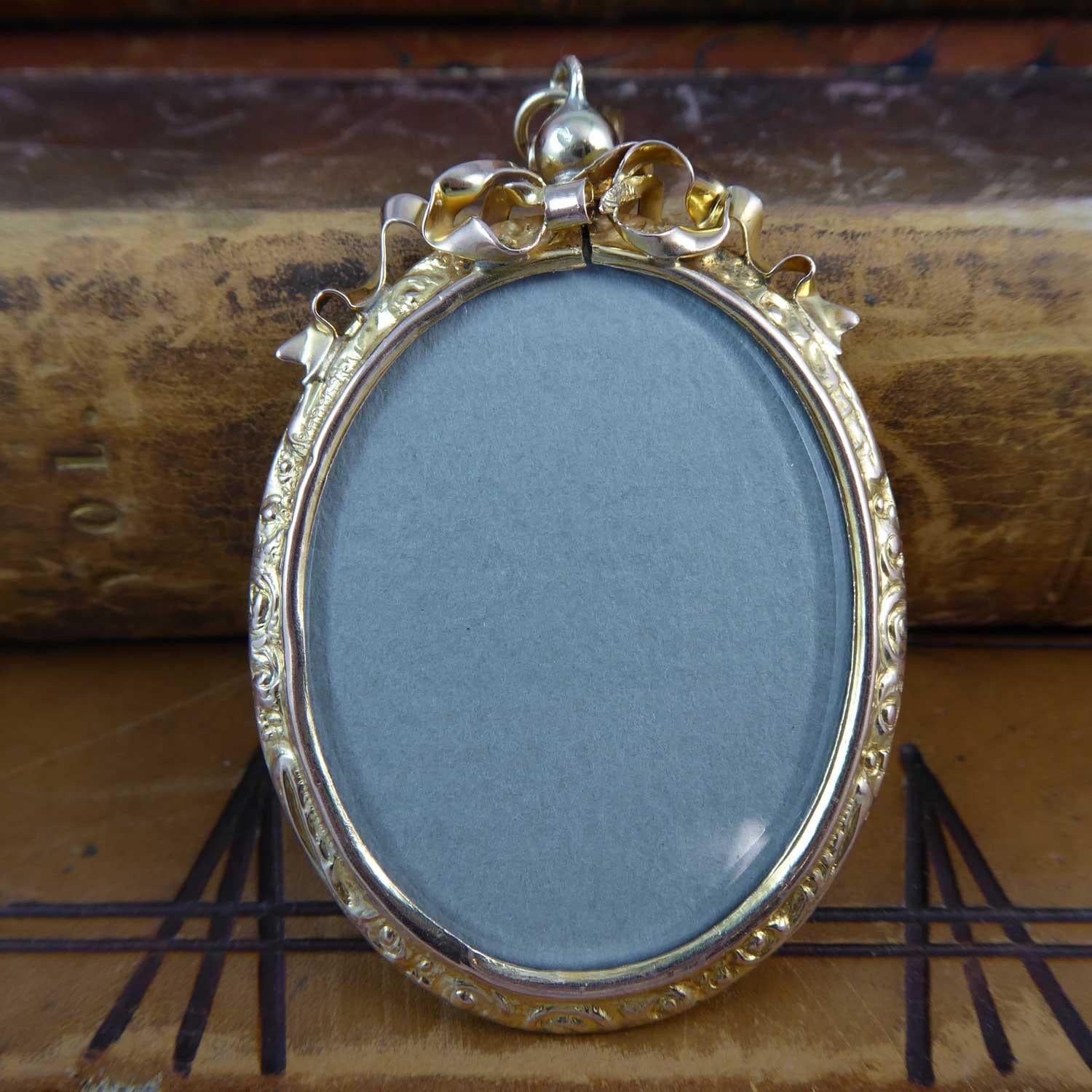 An antique picture locket dating from the Victorian era.  The locket is an elongated oval shape with crystal to both sides.  The edge is a richly carved rococo style engraved pattern with a ribbon bracket style top attached to a bead screw top which