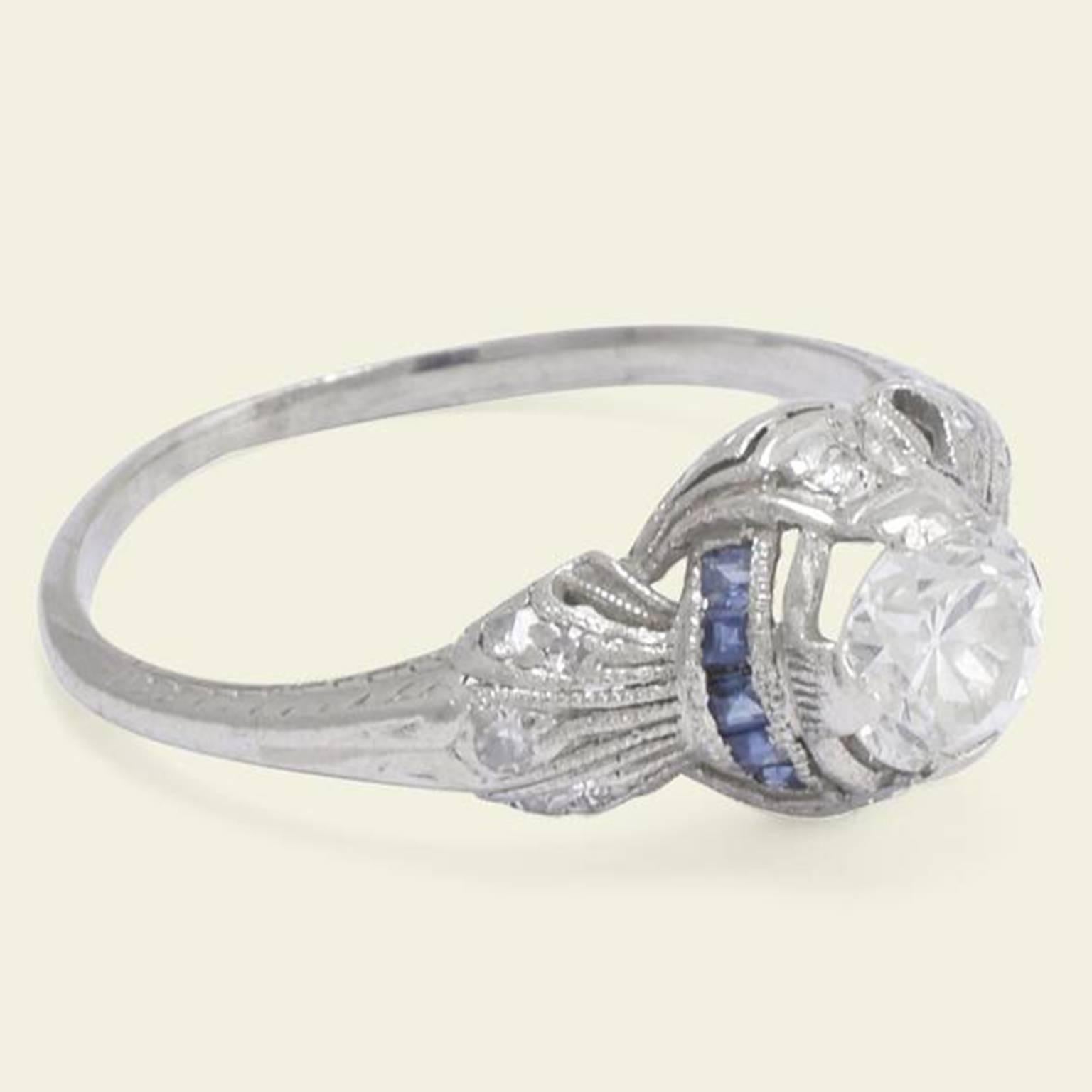 This platinum engagement ring dates to the 1920s. The central .71ct transitional cut diamond (G-H/VS1) surmounts an elaborate setting with diamonds set on the north and south sides and arcs of calibré cut sapphires to the east and west. The fluted