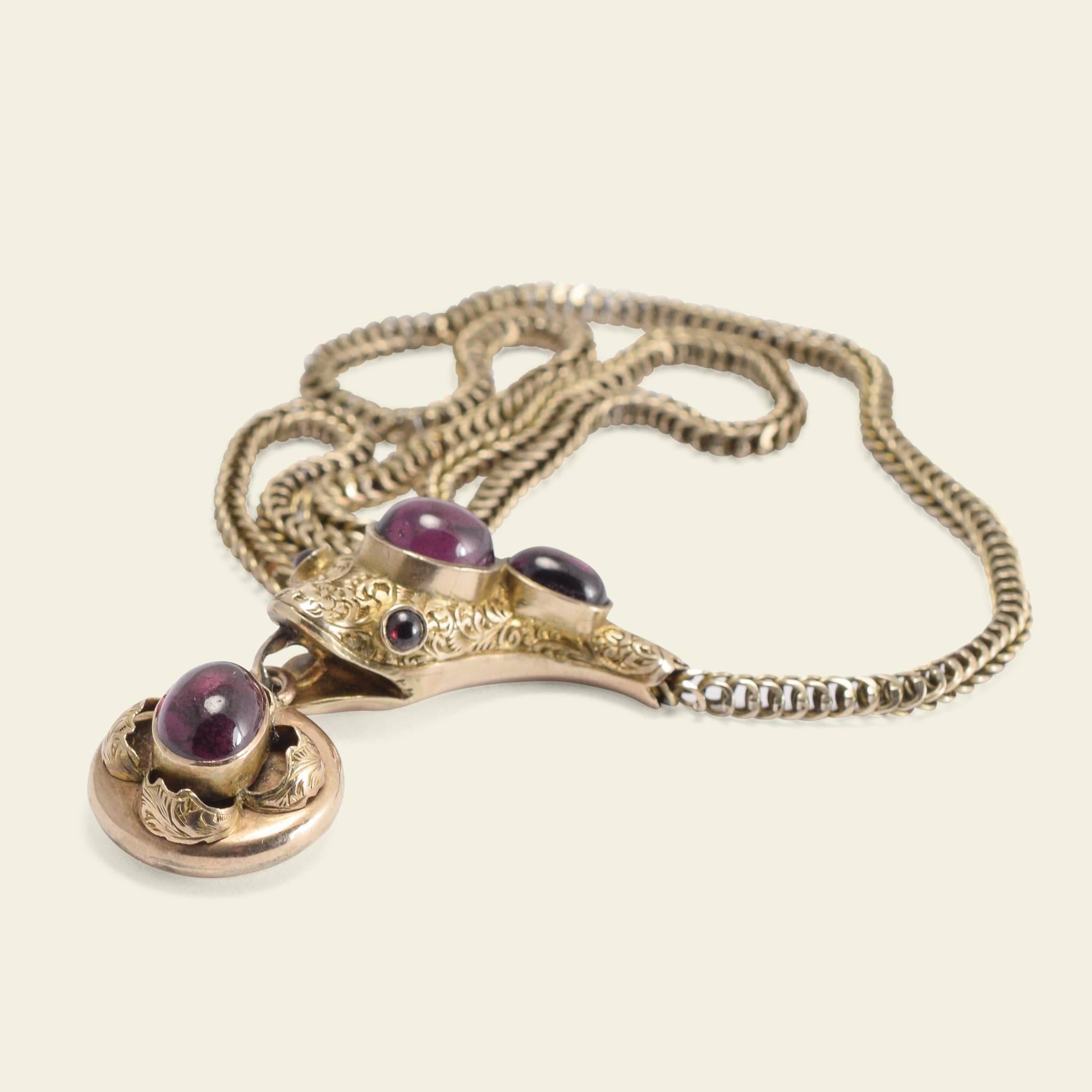 This Victorian era collar necklace is fashioned in the shape of a snake with a lovely linked chain for the body and a chased repoussé head holding a locket in its mouth. The necklace features purplish almandine garnet carbuncles mounted in 9k gold.