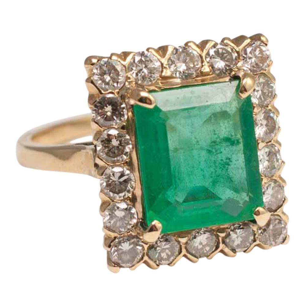 Emerald and diamond ring;  the rectangular emerald is claw set, a vibrant green colour and is surrounded by brilliant cut diamonds; it weighs 3ct with the diamonds totalling 1ct.  Weight 6.6gms.  The diamonds are sitting above a carved gold shank
