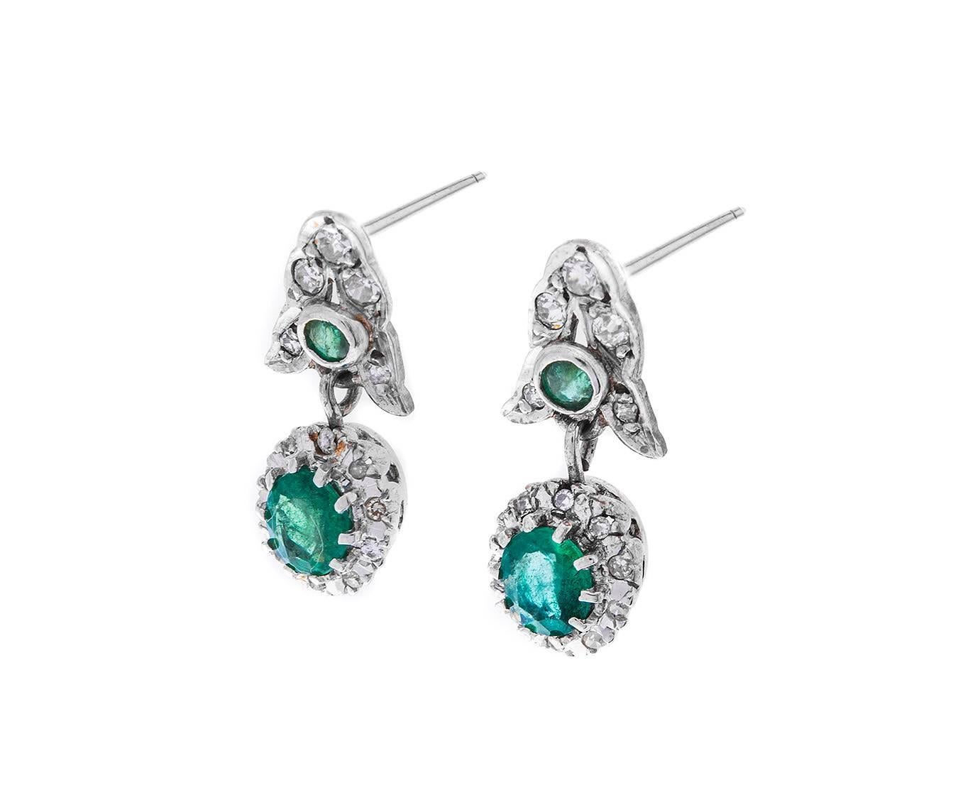 A wonderful pair of antique drop 9ct white gold earrings fashioned in white gold, embellished with diamonds and showcasing beautiful green emeralds. Each earring exhibits a delicious cluster of emerald and diamond suspended from floral motif also