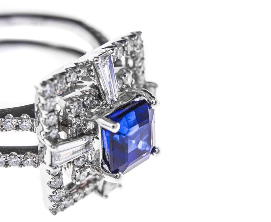 A double square frame of dazzling diamonds with a cross motif of tapered baguette cut diamonds framing a beautiful vibrant blue square emerald cut sapphire and all mounted on a double band of white gold with diamond encrusted shoulders (0.58ct total