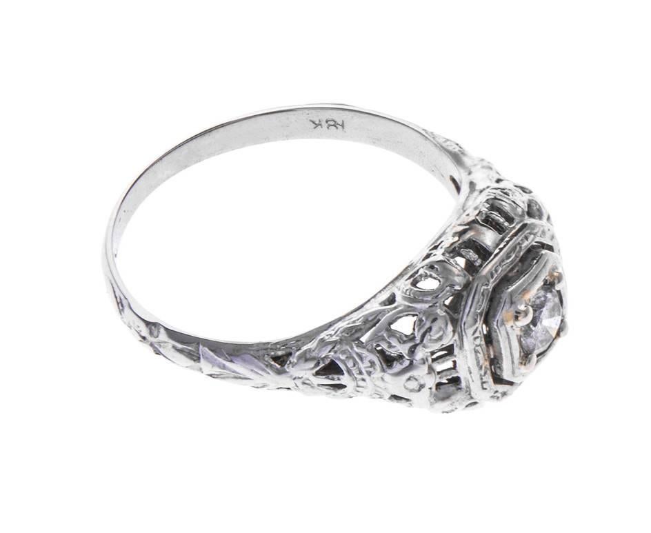 A very Vintage engagement or dress ring, set with a 0.15ct round brilliant cut diamond to intricately decorated halo, shoulders and gallery sides. Designed in white gold, present to someone who has a flair for Vintage flavours.
