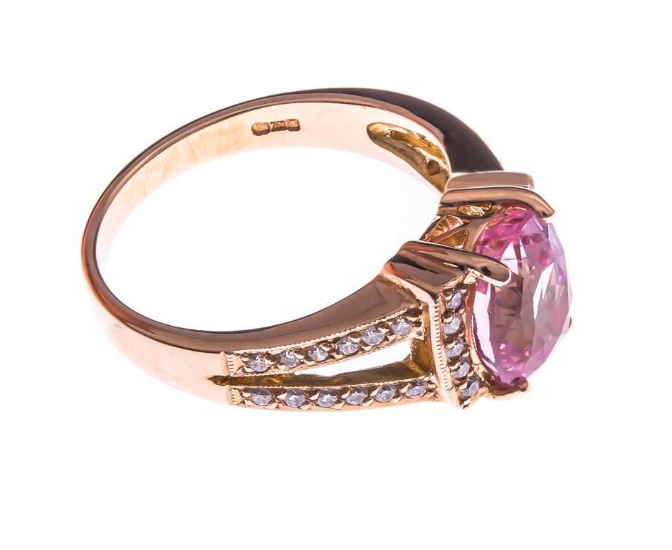 An unusual head turner full of vibrancy and colour. Warm red tones of rose gold encapsulate the bright pink of the fancy sapphire, further enhanced by scintillating round brilliant cut diamonds to the surround and shoulders. A true jewellery