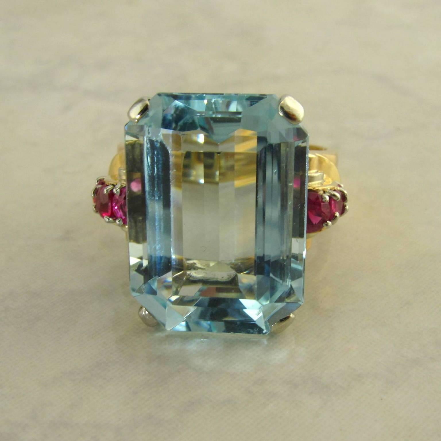 14 karat rose and white gold aquamarine and ruby Retro ring.
One center emerald cut aquamarine set in white gold weighing approximately 13.30 carats, accented with six round prong set synthetic rubies weighing approximately .60 carat total. The