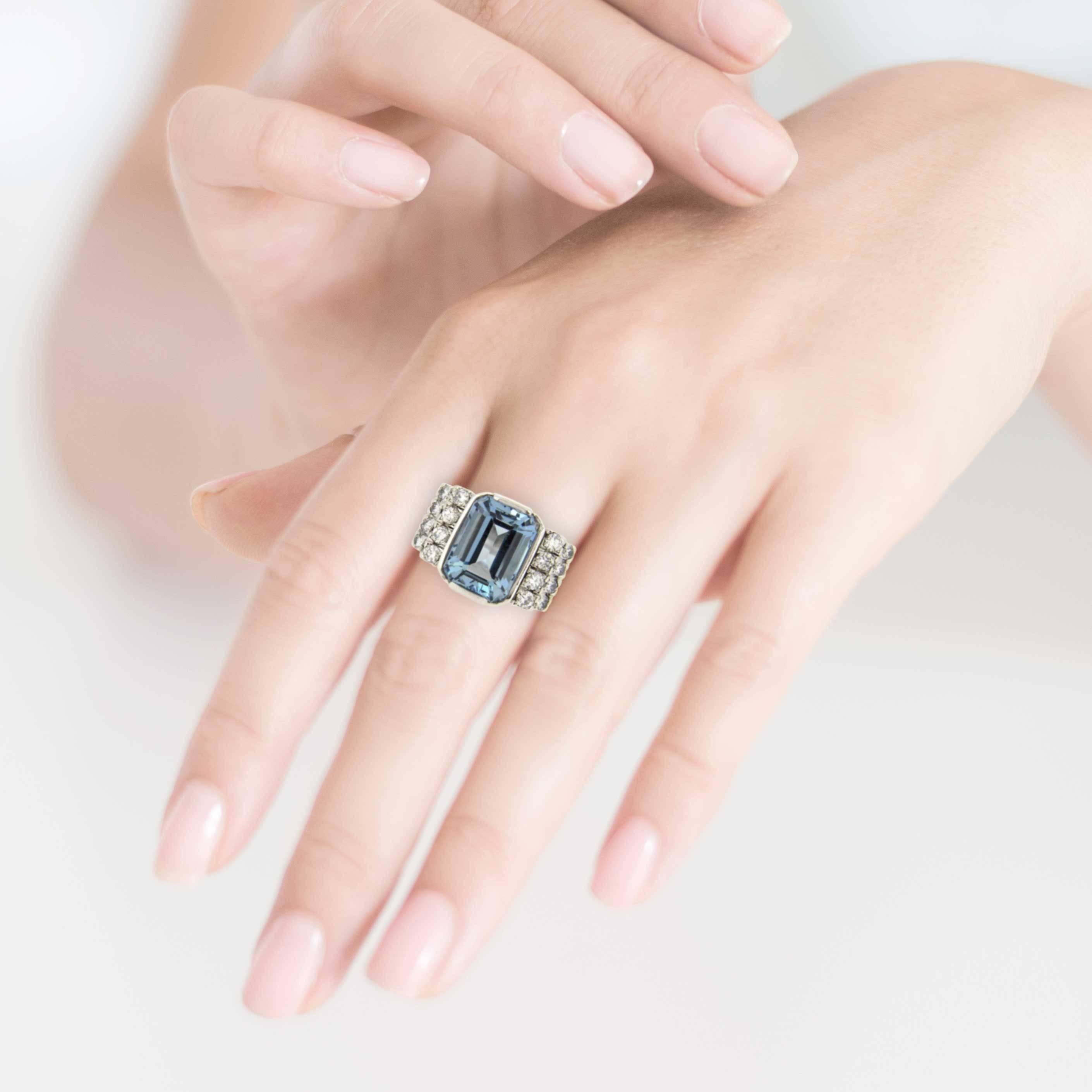 An elegant handmade 18 karat white gold centred with one emerald cut medium blue coloured natural aquamarine; flanked by double stepped shoulders set with a total of sixteen claw set brilliant cut diamonds. 
This classical design certainly makes a