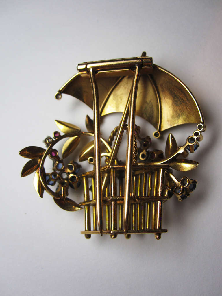 Retro yellow gold brooch of a stylised flower basket with an umbrella. Flowers gem set in Ruby, Sapphire and Diamond. In total, 9 round cut diamonds with an approximate weight of 0.50ct, 10 round cut sapphires and  11 round cut Rubies. Circa 1940s.