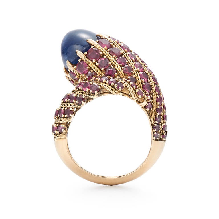 A natural unheated cabochon sapphire weighing approximately 12.75cts, with gemological report, is accented with round rubies totaling approximately 3.65cts in an 18 karat yellow gold mounting signed Marchak Paris, numbered. The ring measures
