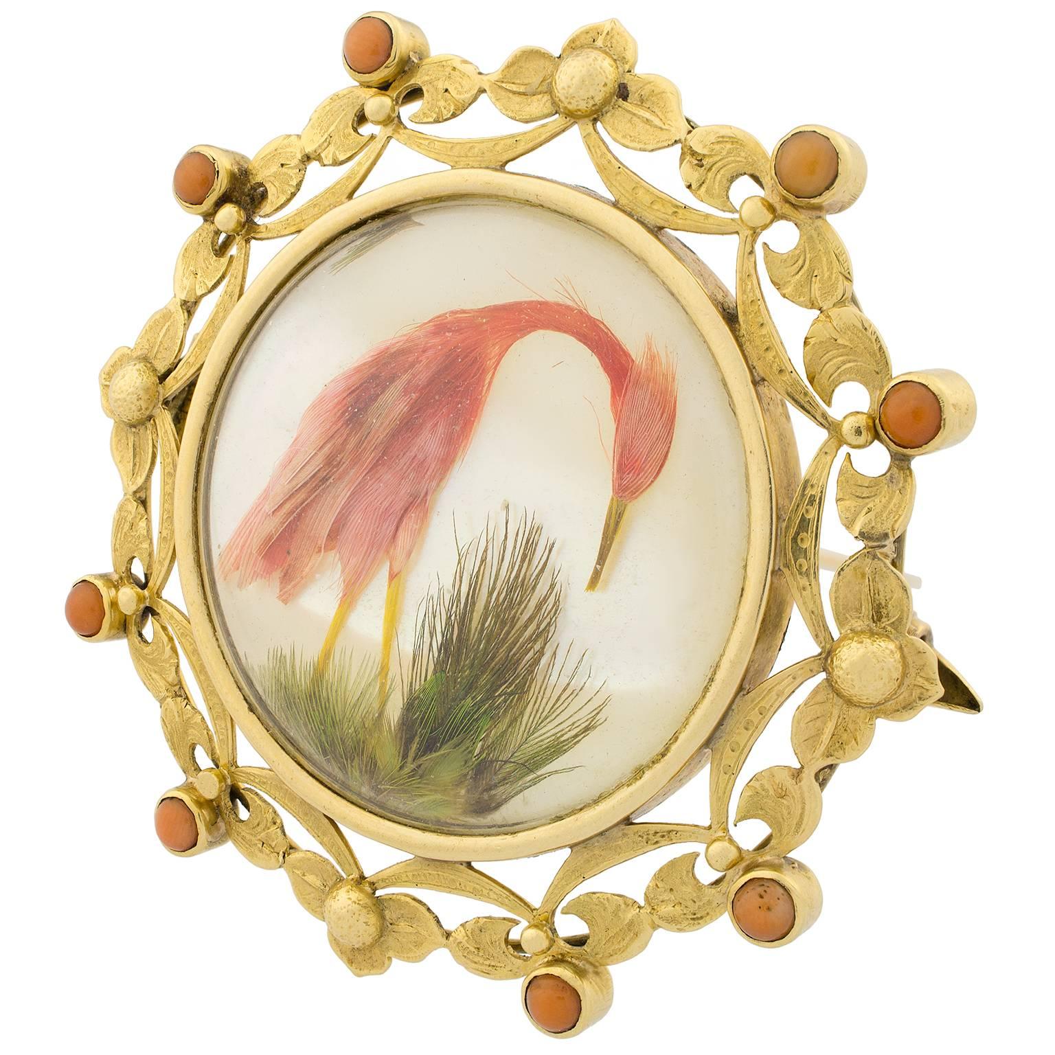 Victorian brooch in gold depicting a heron with details in coral, feathers and mother of pearl, inside a textured foliate frame.