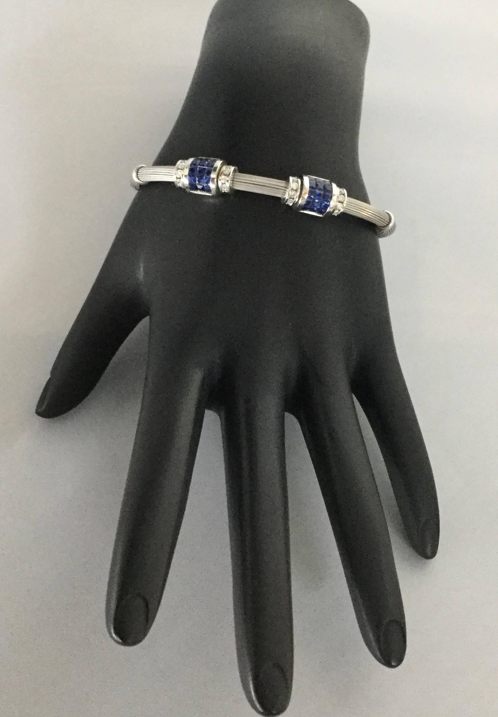 18K white gold flexible cable cuff bracelet with two sapphire and diamond stations. The bracelet contains 30 princess cut invisible set sapphires weighing combined 0.75 carat and 16 round diamonds weighing combined 0.38 carat.
