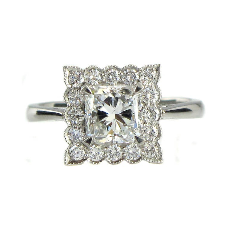 A platinum diamond ring, comprising a 1.03 carat cut cornered rectangular modified brilliant cut diamond, certified by GIA as being E colour, SI2 clarity. Set within a classic milgrain set round brilliant cut diamond surround.

This ring has been