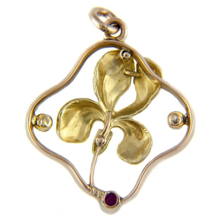 An Art Nouveau gold pendant in the form of an iris set within a natural pearl and ruby surround in the style of Krementz. Circa 1910. Stamped 9ct. Tests as 9ct gold.

Approximate size 3cm x 3.5cm. The enamel work is in perfect condition.