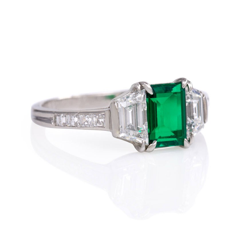 An Art Deco emerald and diamond ring, the center rectangular emerald flanked on both sides by a trapezoidal diamond and four square-cut diamonds, in platinum.  Tiffany & Co.; original Tiffany box.  Engraved D.M.B. 11-20-35.  Atw emerald 0.66 ct.,