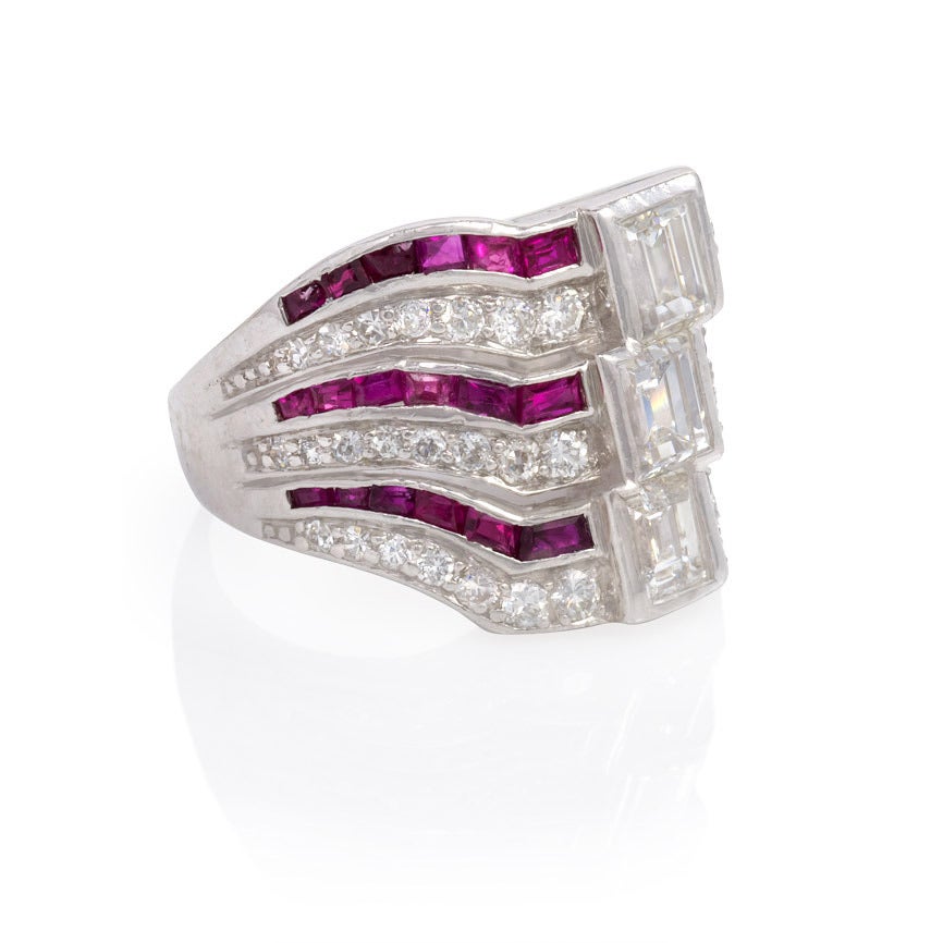 An Art Deco three-stone emerald-cut diamond ring of stacked, wave-form design with bands of calibré rubies, in platinum.