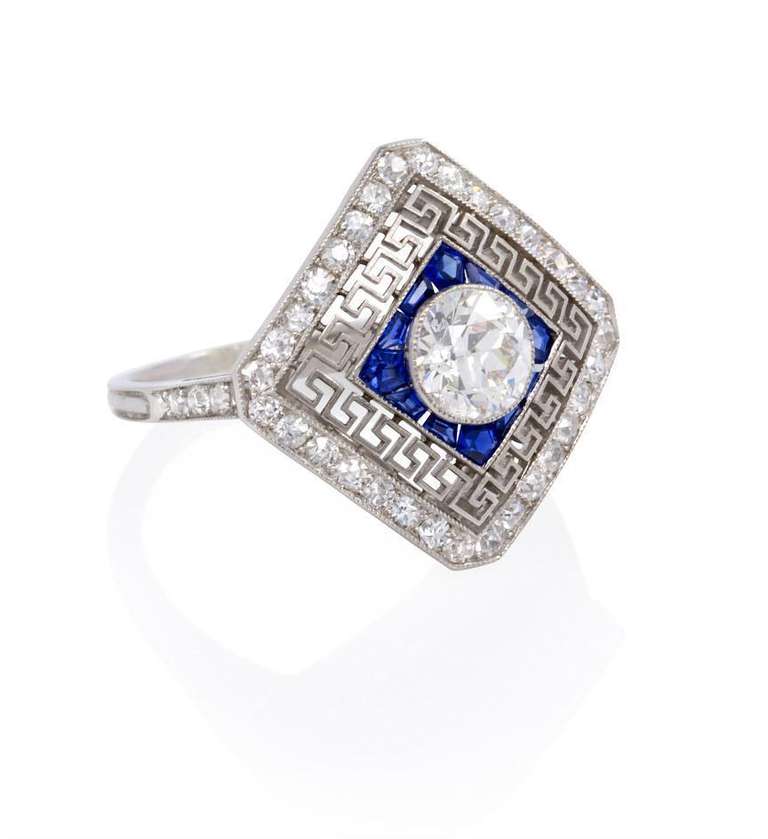 An Edwardian diamond ring with a central old European cut diamond surrounded by calibre cut sapphires, a filigree Greek key design, and a diamond-set border, in platinum.  Atw center stone 0.75 ct., J-K color, SI1 clarity