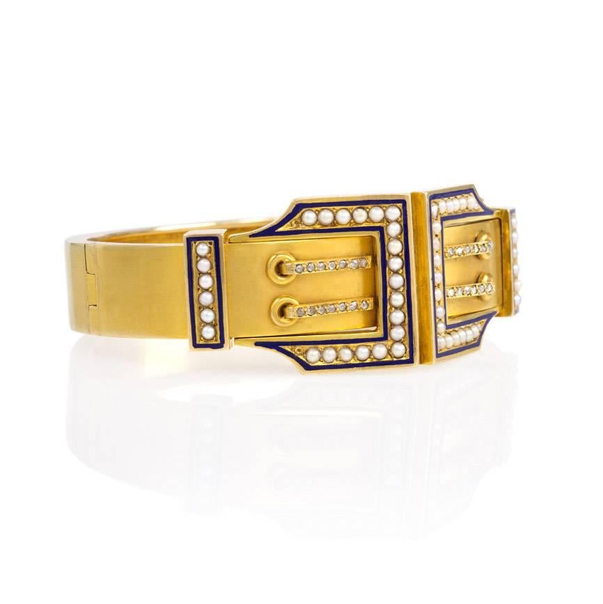 An antique gold bangle bracelet with a double-buckle motif comprised of half-pearls and diamonds with blue enamel detailing, in 14k.  Inner circumference: 6.5