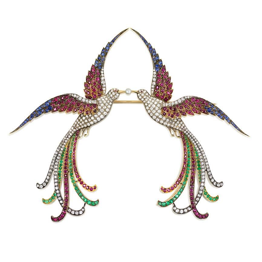 A superb antique brooch in the form of a pair of birds of paradise with diamond bodies and emerald, sapphire and ruby plumage and joined by a pearl at their beaks. The birds separate to form two separate brooches, in sterling silver and 18k gold. 