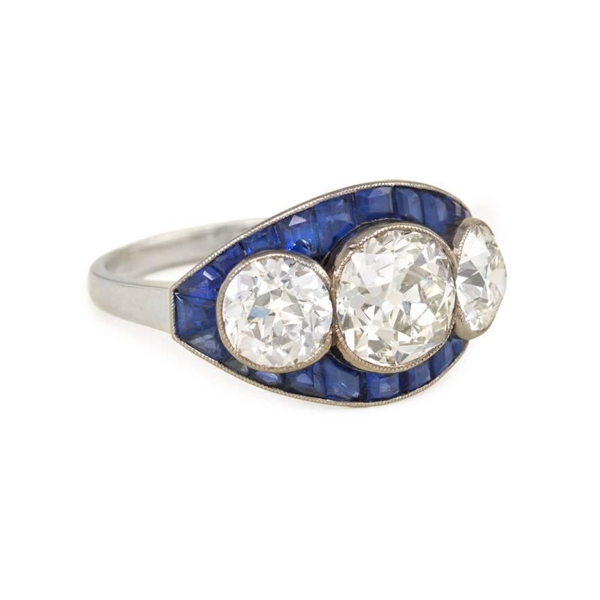 An Art Deco three stone cushion-cut diamond ring with a calibré sapphire surround, in 18k gold and platinum.  France.  Atw diamonds 3.21 cts.