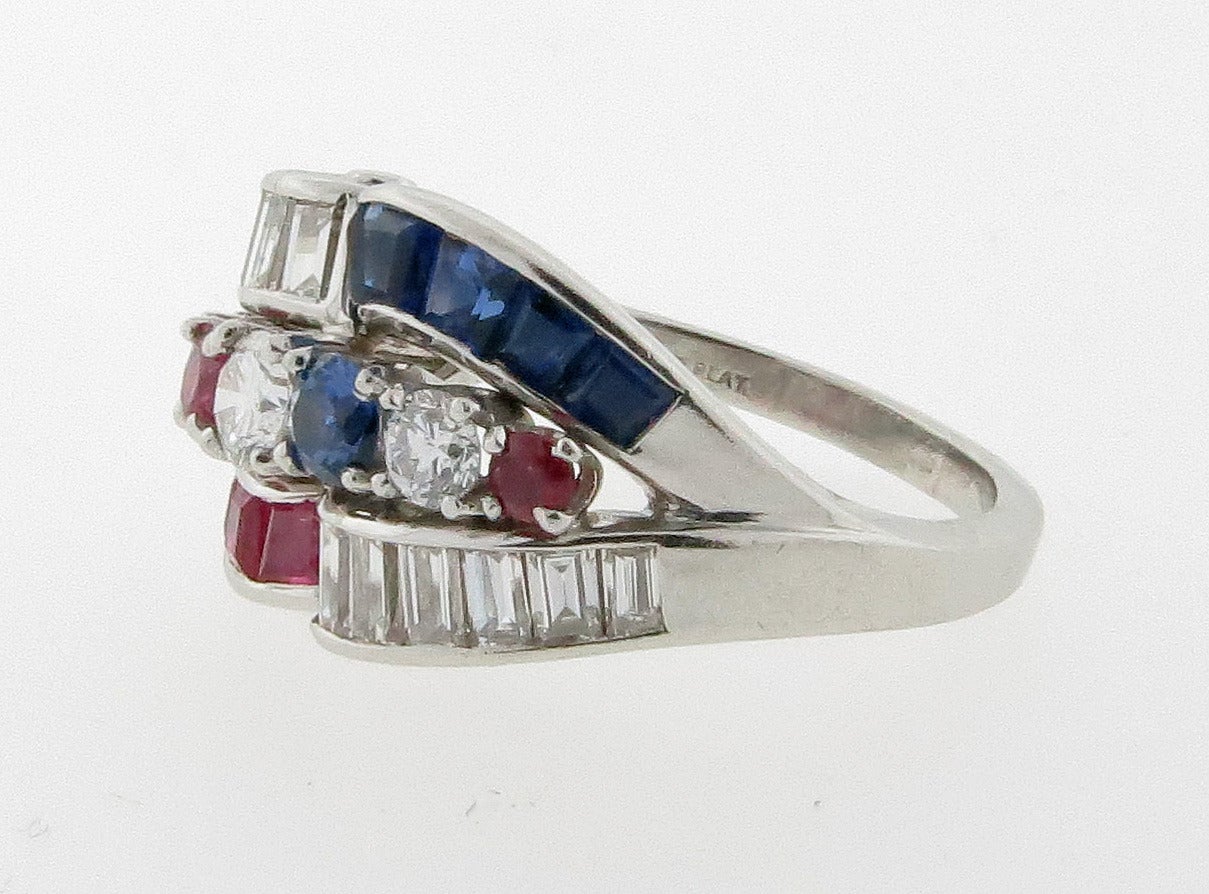 Handmade Bi-Centennial  platinum mount crossover design ring. Prong set across the top with two rubies,  sapphires, and three round brilliant cut diamonds.  Alternating flanked by five channel set rubies and sapphires, and channel set by 14 baguette