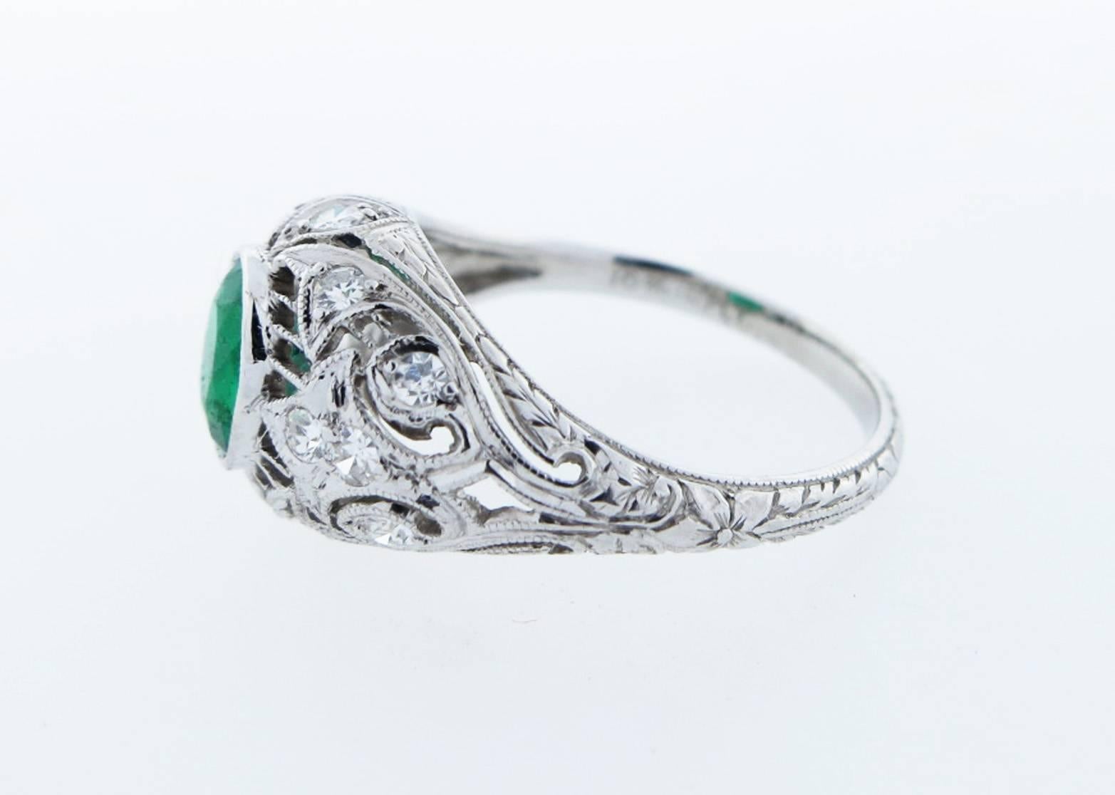 Unique open work platinum ring circa 1920. Bezel set with a center oval faceted natural emerald measuring 6.3 mm. x 5.2 mm. weighing approx .70cts. Bead set in an exquisitely engraved mount with 14 round diamond accents totaling approx .20cts.  size
