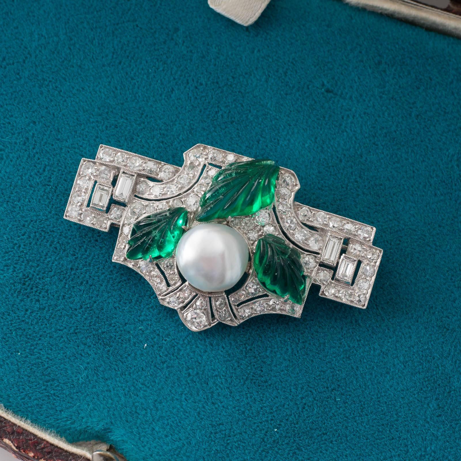 An Art Deco platinum brooch featuring a 4.49ct button shaped natural pearl set within a pierced and diamond set plaque with three large emeralds carved into the shape of leaves arranged around it with a pin roller catch and jump ring to the reverse.