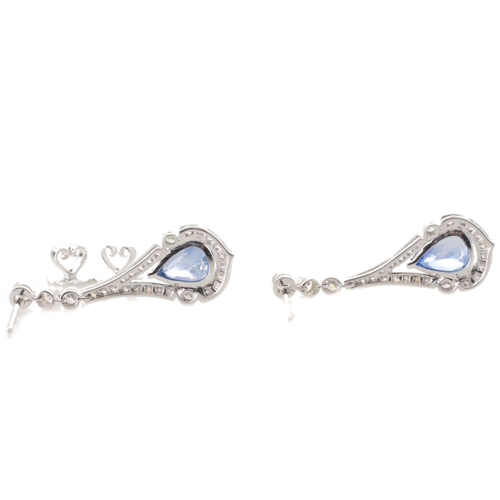 A pair of 18ct white gold drop earrings each with a pear shaped sapphire GIA certified as Sri Lankan in an arabesque styled surround which is grain set with brilliant cut diamonds and bezel set with a diamond to either side of the sapphire all