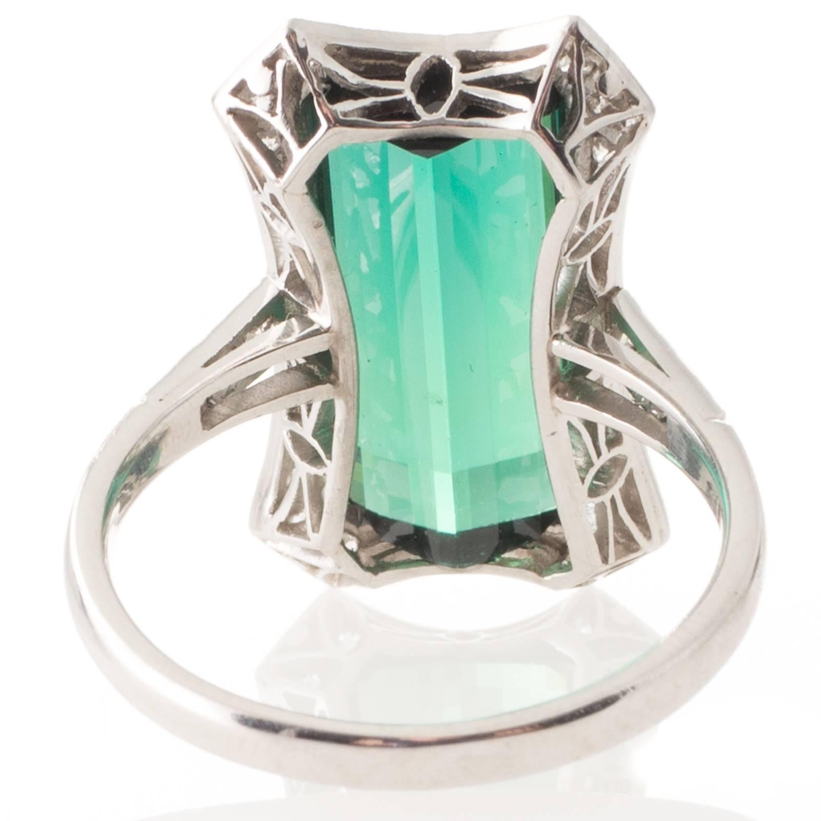 An 18ct white gold  ring, bezel set with a rectangular cut green tourmaline of 7.09ct, in a frame of twelve grain set early brilliant cut diamonds above a pierced gallery to upswept grain set diamond shoulders and a plain polished band. 
Total