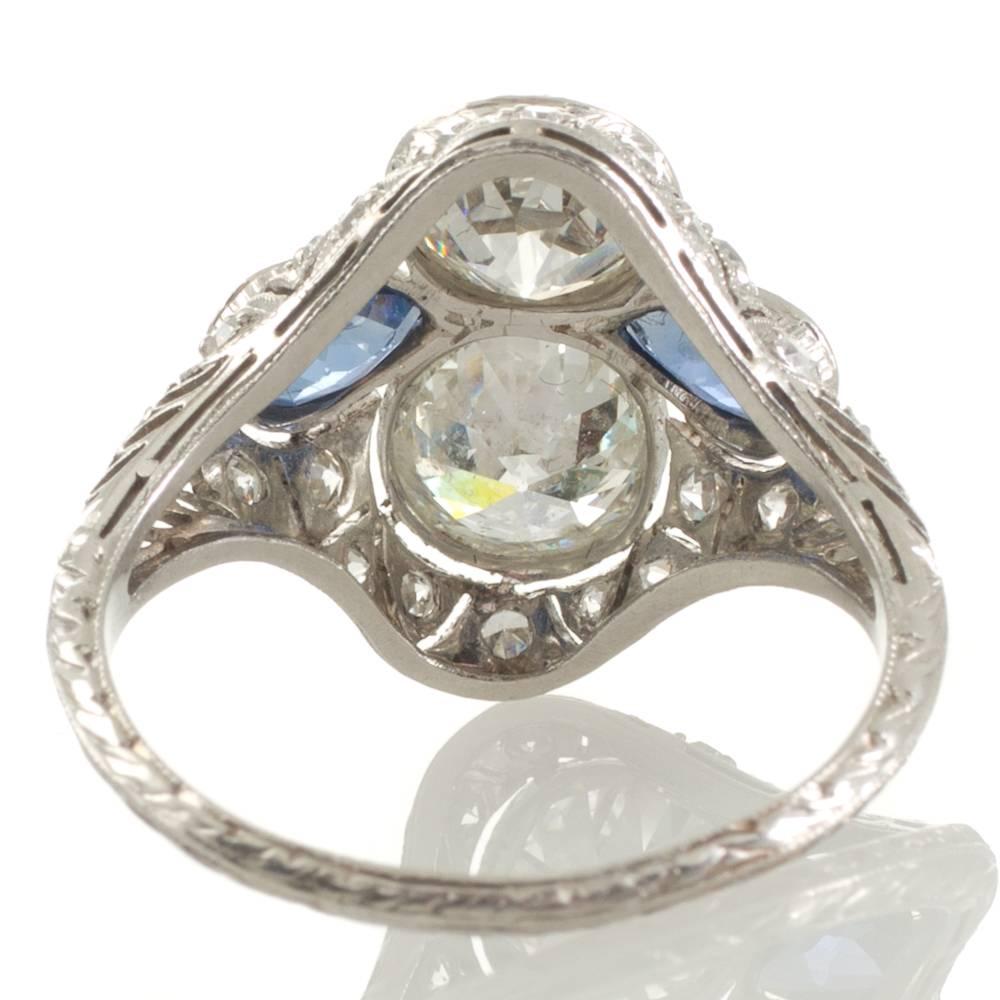 An antique platinum plaque ring with two early transition cut diamonds of total weight 2.60ct graded as colour J-L and clarity P1-2 in engraved raised bezel settings with millegrain edges and two pear shaped blue sapphires in matching settings all