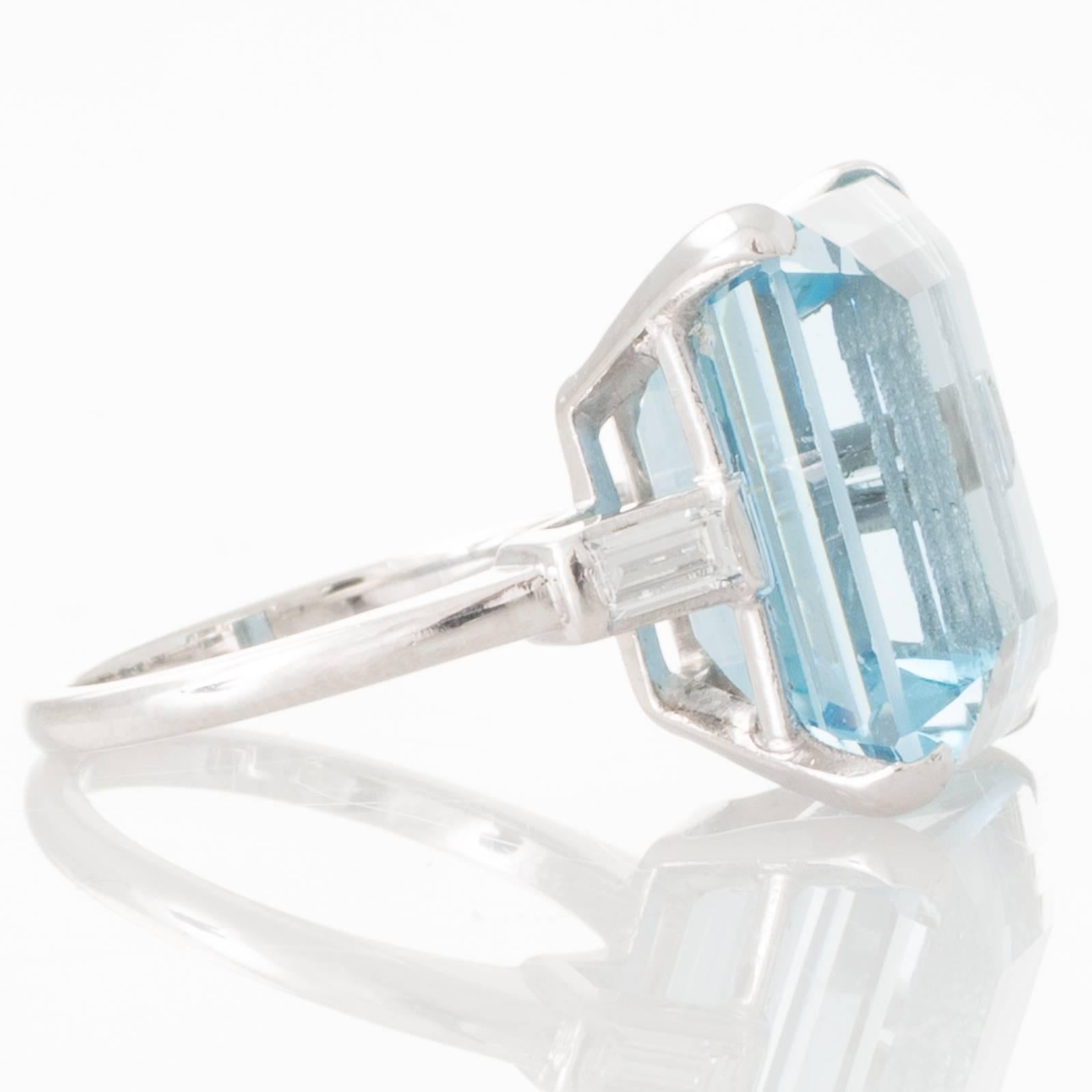 Platinum ring set in four corner claws with an estimated 12.45ct step cut aquamarine in a double railed box mount, and to either side a baguete cut diamond in the sloping shoulders and a plain polished band.
Total Estimated Aquamarine Weight: