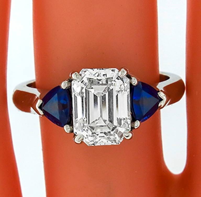 This platinum ring centers a sparkling GIA certified emerald cut diamond that weighs 1.94 carat and is graded H color with VS1 clarity. The center stone is flanked by two vivid blue trilliant cut sapphires weighing approximately 0.80 carat. 
The