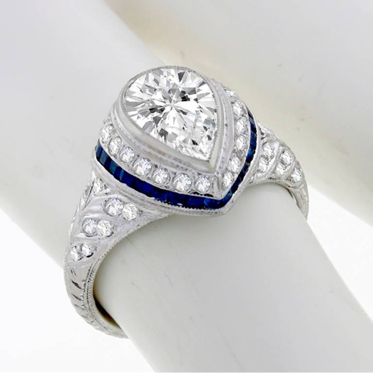 This platinum engagement ring from the Art Deco era, centers a sparkling GIA certified pear shaped diamond that weighs 1.18ct. and is graded F color with VS2 clarity. The center diamond is accentuated by vivid blue square cut sapphires weighing