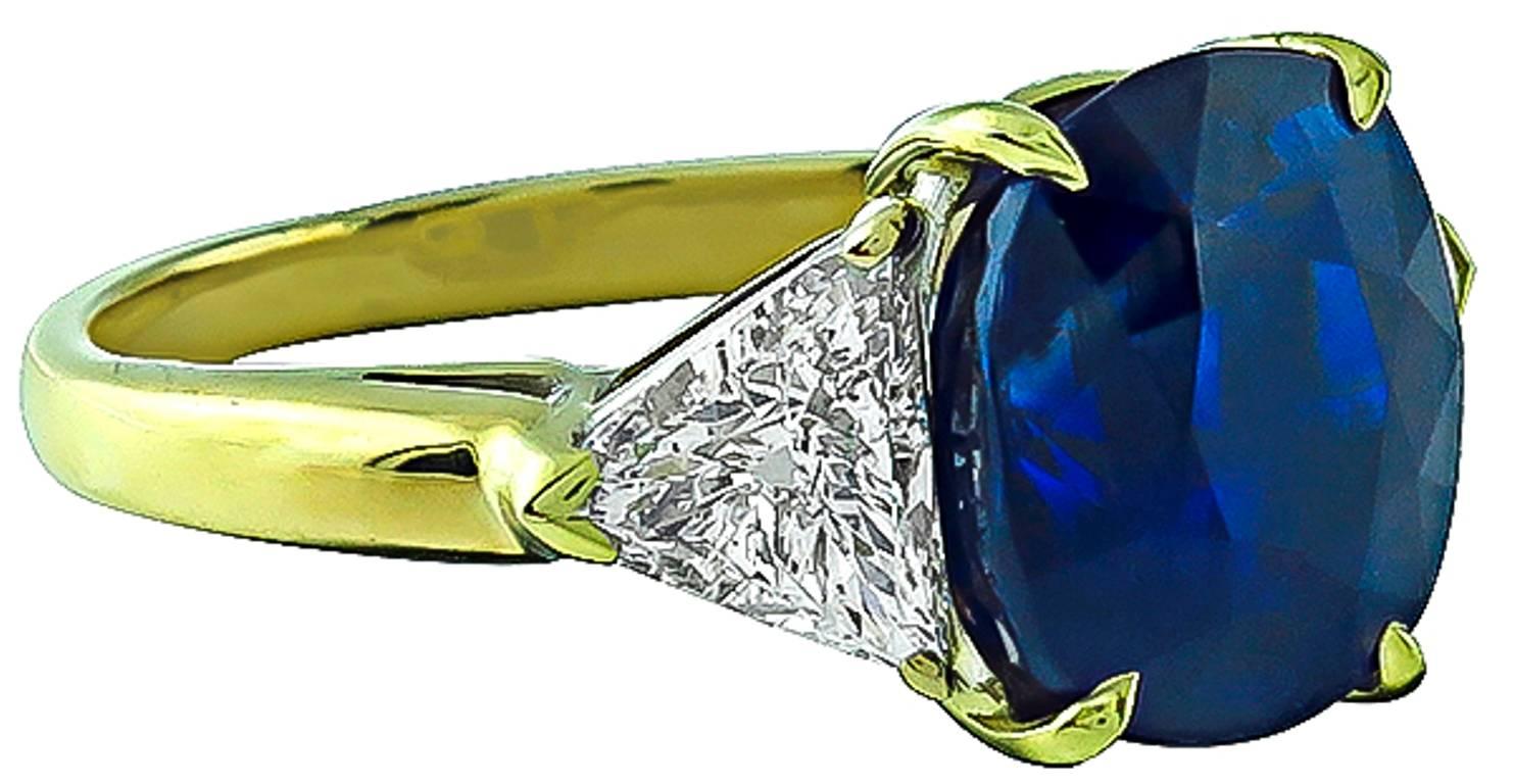Made of 18k yellow gold, this ring is centered with a vivid blue cushion cut Ceylon sapphire that weighs 5.28ct. The center stone is flanked by sparkling trilliant cut diamonds that weigh approximately 1.00ct. and are graded G-H color with VS2