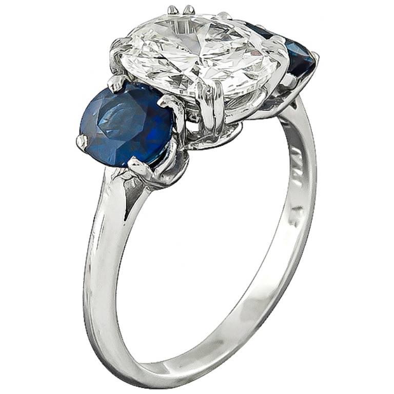 This stunning platinum ring is centered with sparkling oval cut diamond that weighs 2.12ct. graded J-K color with VS1 clarity. The center diamond is flanked by high quality oval cut sapphires that weigh 1.88ct. The top of the ring measures 9mm by