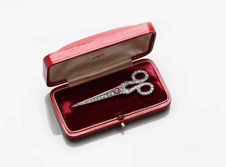 Shaped like a pair of scissors, this silver and gold brooch is encrusted with rose-cut diamonds and a central ruby.

With fitted case.

Unmarked