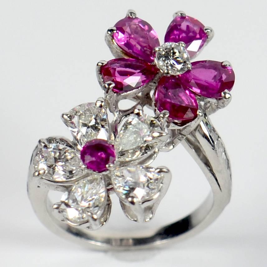 A fine ruby and diamond crossover ring in platinum set with two flowerhead terminals – one with five Burma ruby pear-shaped petals to a round brilliant cut diamond centre, the other with five diamond petals to a Burma ruby center.  The shoulders are