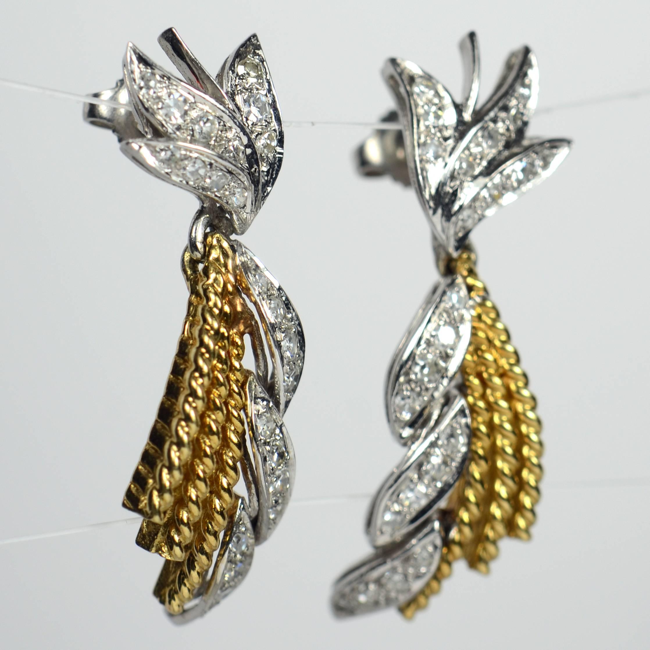 A pair of drop earrings in 18 carat white and yellow gold set with diamonds.  Each has a diamond set white gold stud designed as a spray of three leaves with a pendant drop of three curved twisted yellow wires next to a curved and twisted ribbon of