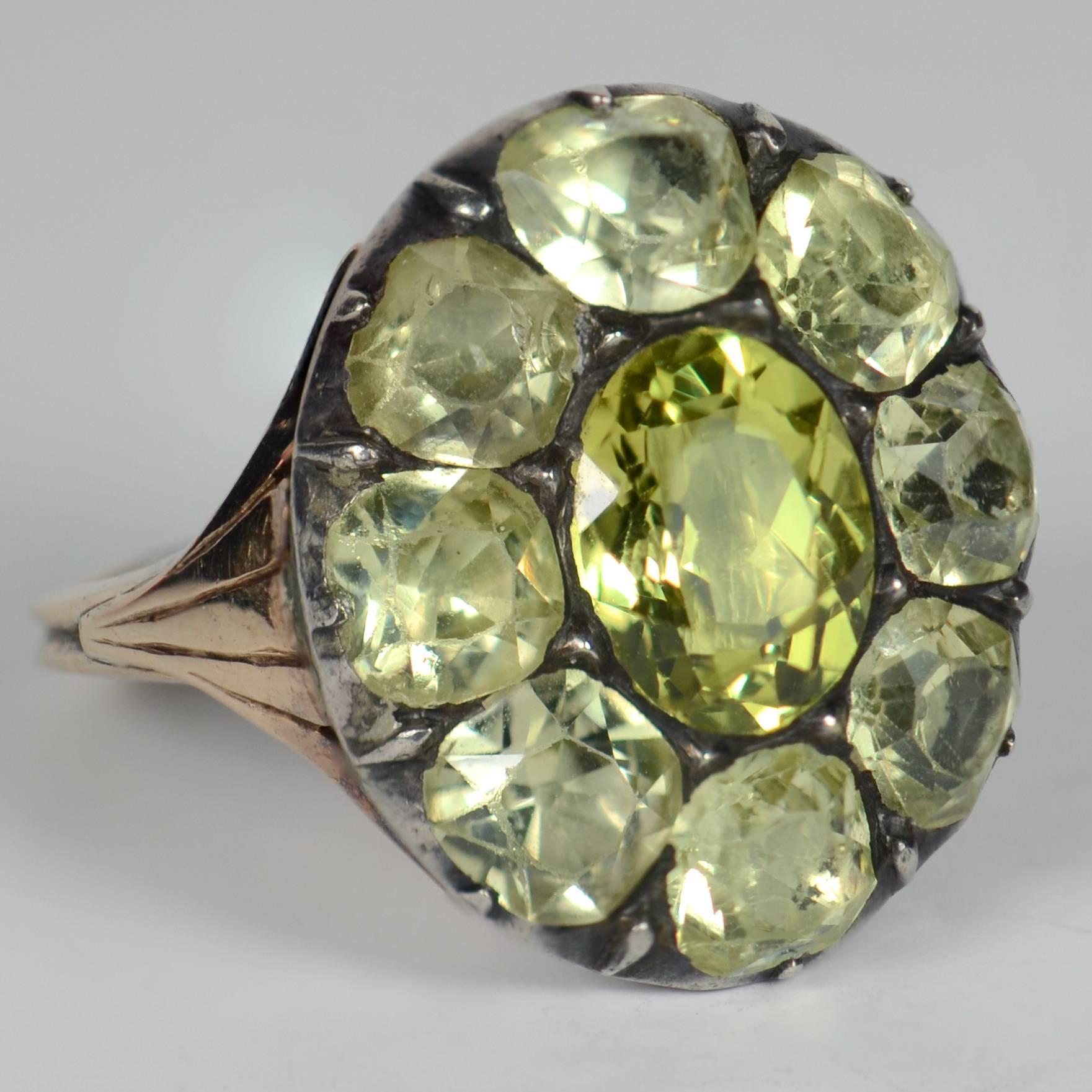This antique Portuguese cluster ring has a large central, intensely green, chrysoberyl with a surround of eight slightly smaller chrysoberyls. The stones are mounted in silver-topped gold with a foliate design to the shoulders leading to a slim