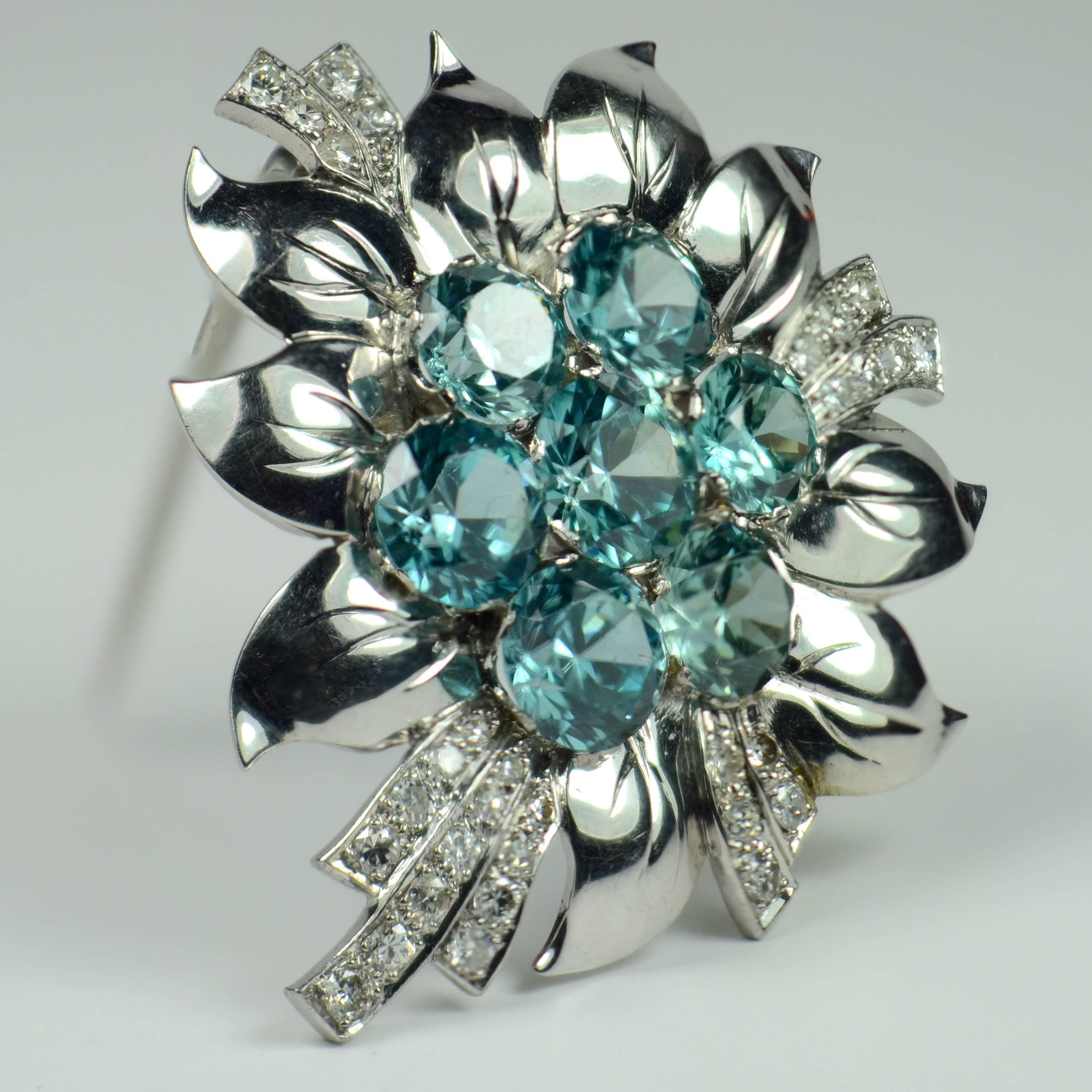 A beautifully made brooch in the form of a flower with a blue zircon heart surrounded by curving platinum leaf shapes with engraved veins and diamond set curved tendrils.

The centre of this brooch is set with seven zircons with a fine sky blue