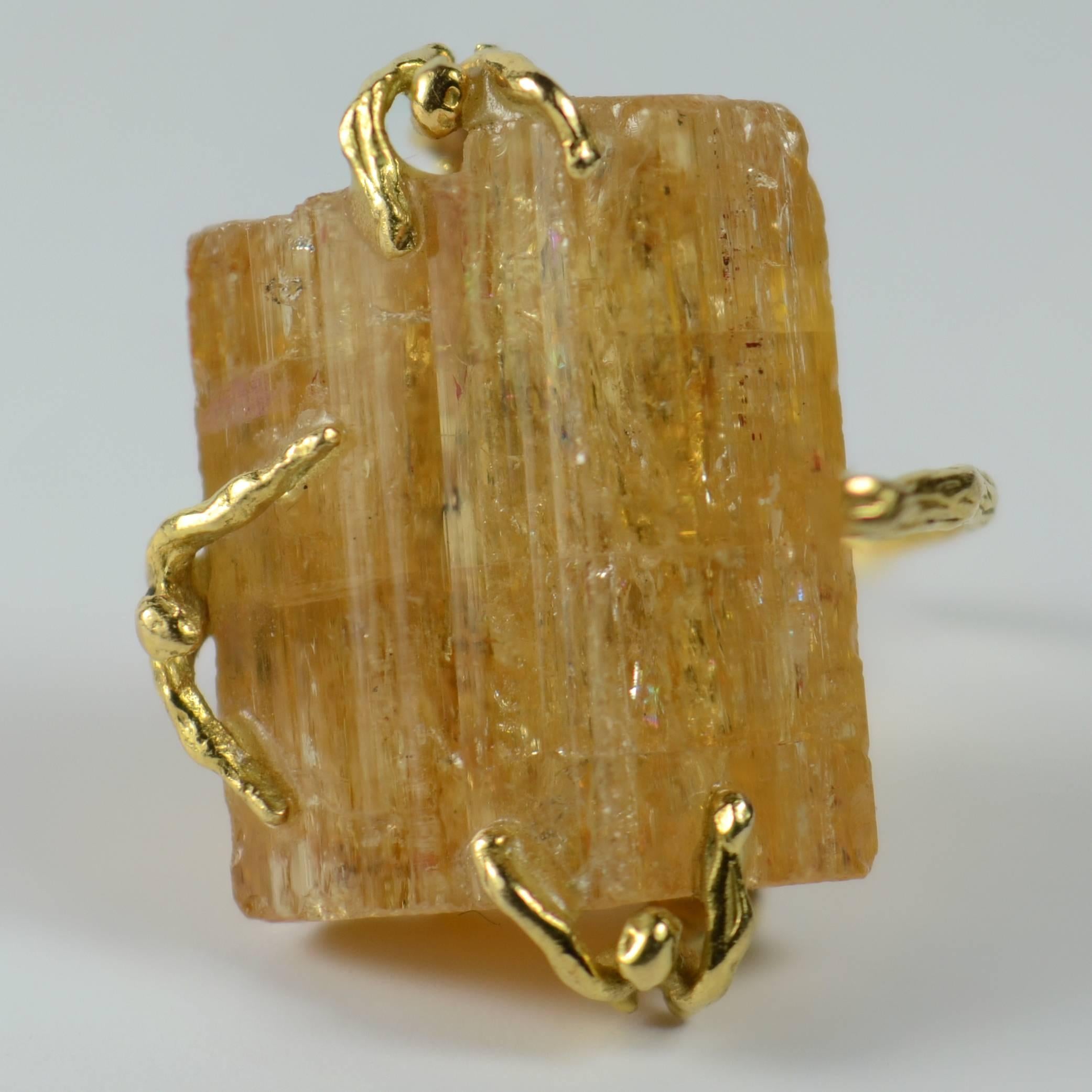 An artist-made ring depicting three figures clinging to a crystal of Brazilian orange topaz. The figures are fashioned from 18 carat gold wire and are standing on a branch, which forms the shank of this ring.  The topaz crystal measures 2.2cm x