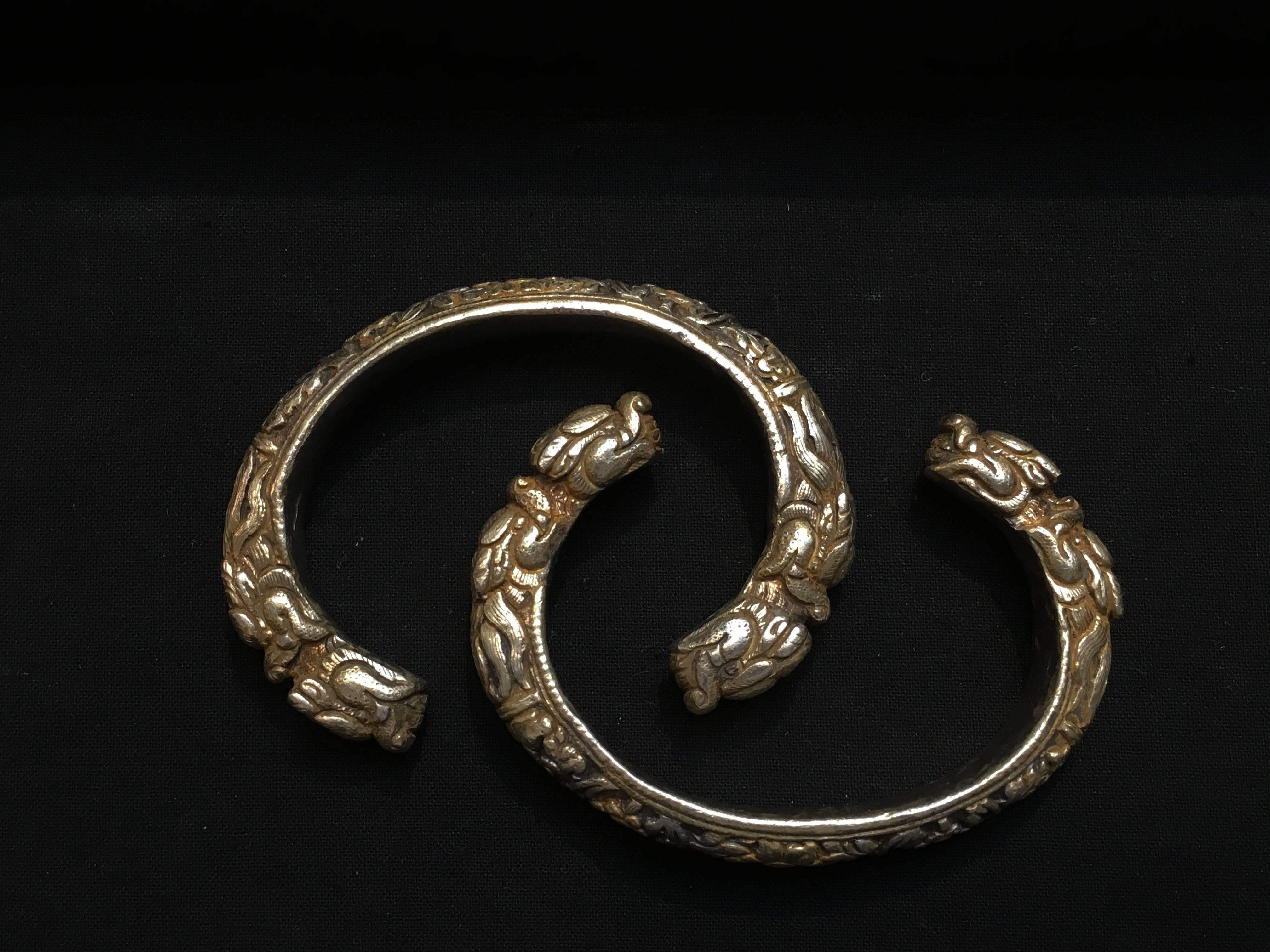 A heavy pair of cast silver and parcel gilt bangle bracelets from Bhutan, mid 20th century. Of traditional Himalayan style, the c-shaped bangles decorated with a deeply carved floral and foliate band terminating in a pair of unusual double makara