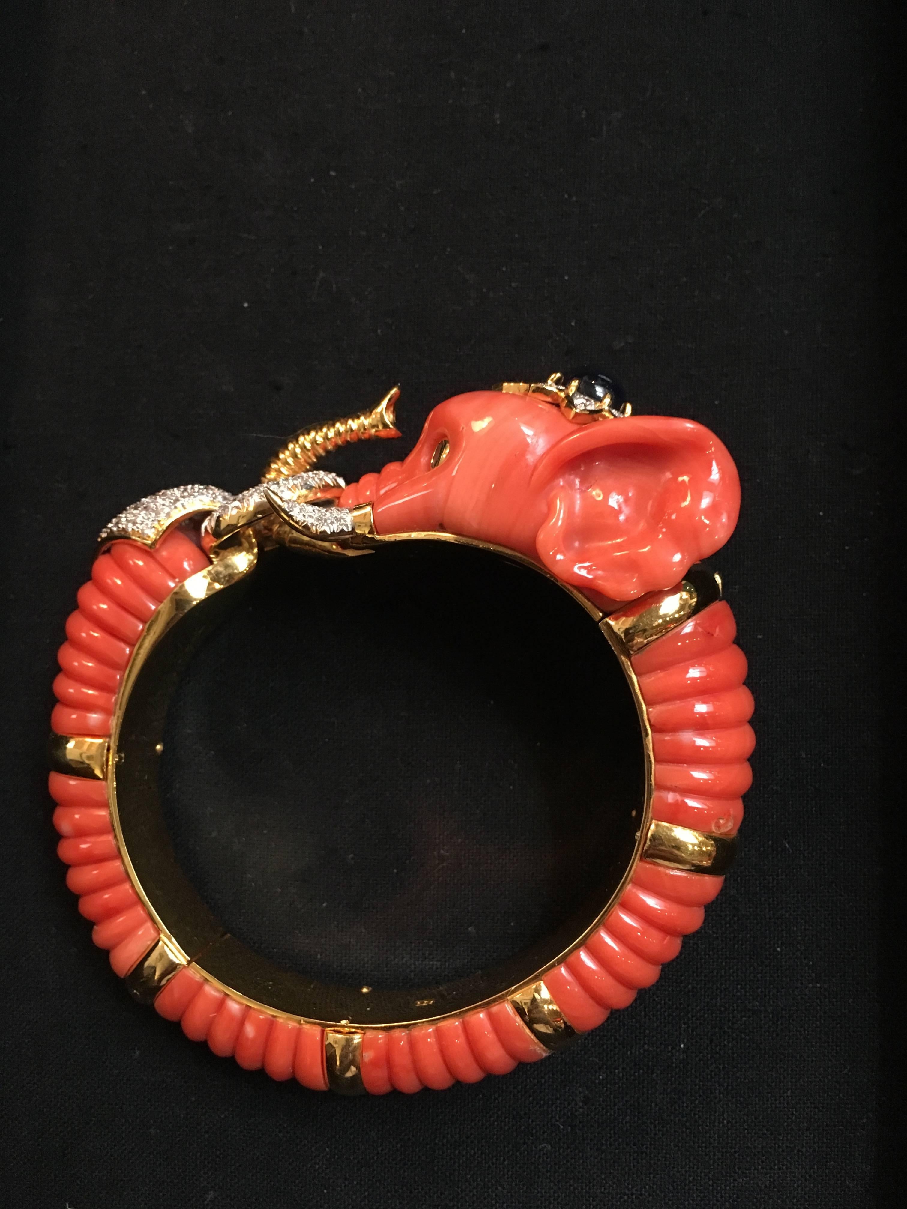 A stunning and charming  animal bracelet in form of a carved coral elephant. The coral of an overall even salmon color. The head set with emeralds for eyes, and a large sapphire to the top surrounded by diamonds. The gold tusks also set with