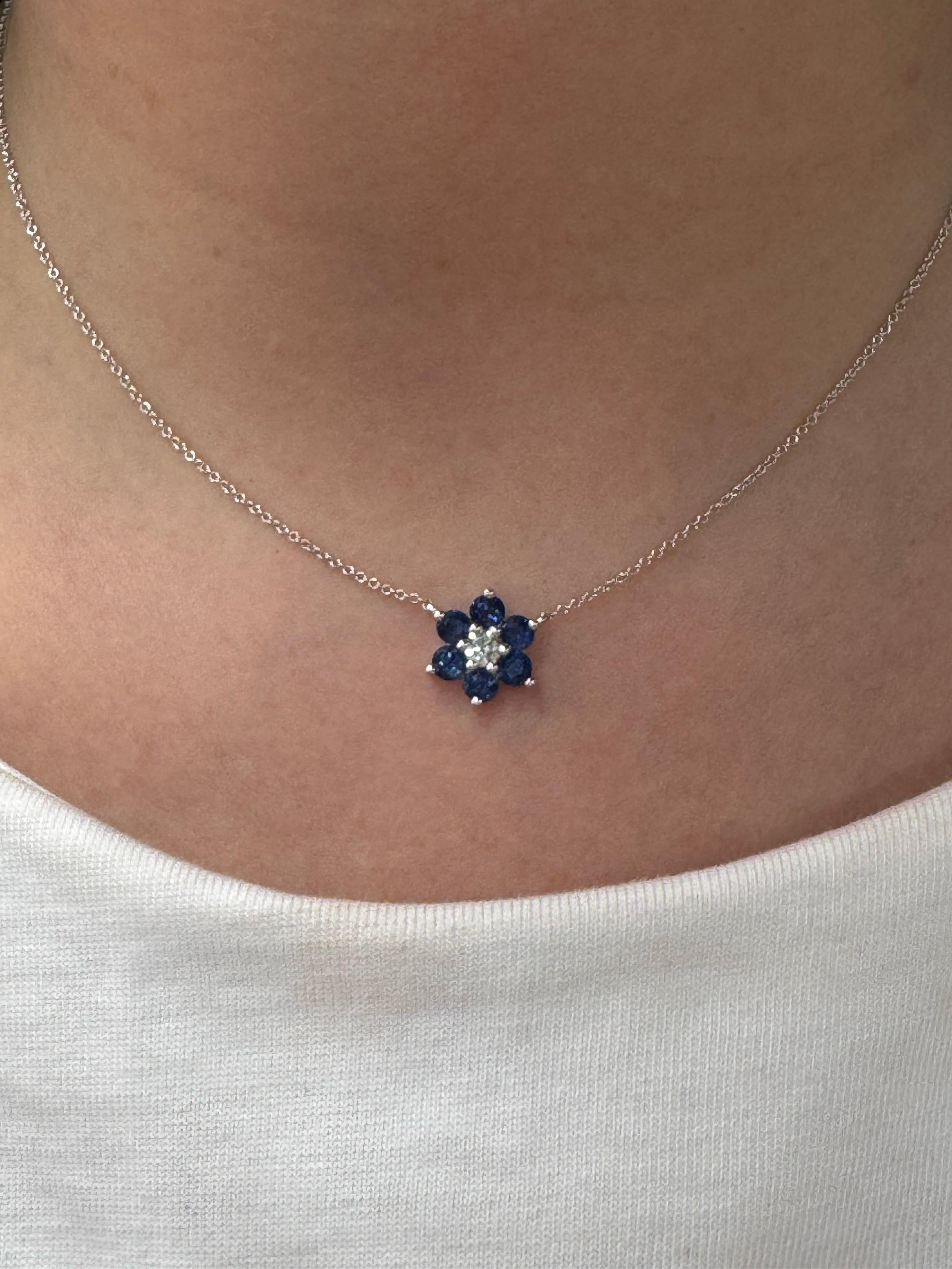 Diese elegante Halskette aus 14-karätigem Weißgold zeichnet sich durch einen charmanten blumenförmigen Anhänger aus. In seiner Mitte funkelt ein runder Brillant mit einem Gewicht von 0,18 Karat, umgeben von sechs blauen Saphiren mit einem