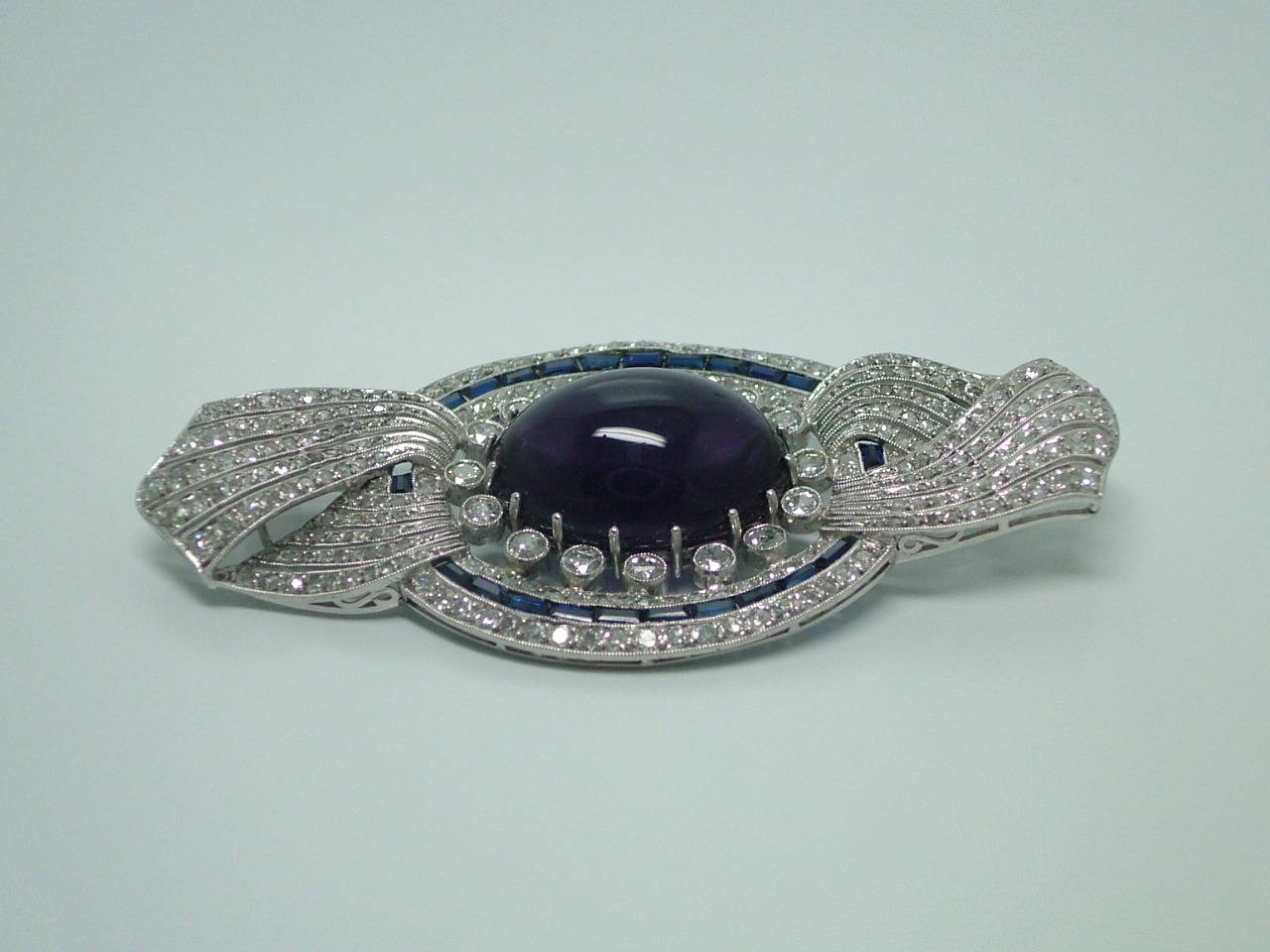The oval cabochon Amethyst measuring 20x14mm surround of delicate calibré rectangular blue sapphires and old-mine diamonds is partial surround by a rose cut diamonds set ribbon.

STONES ESTIMATED WEIGHTS: Sapphire: 2.00ct. Diamond: 4.50ct.